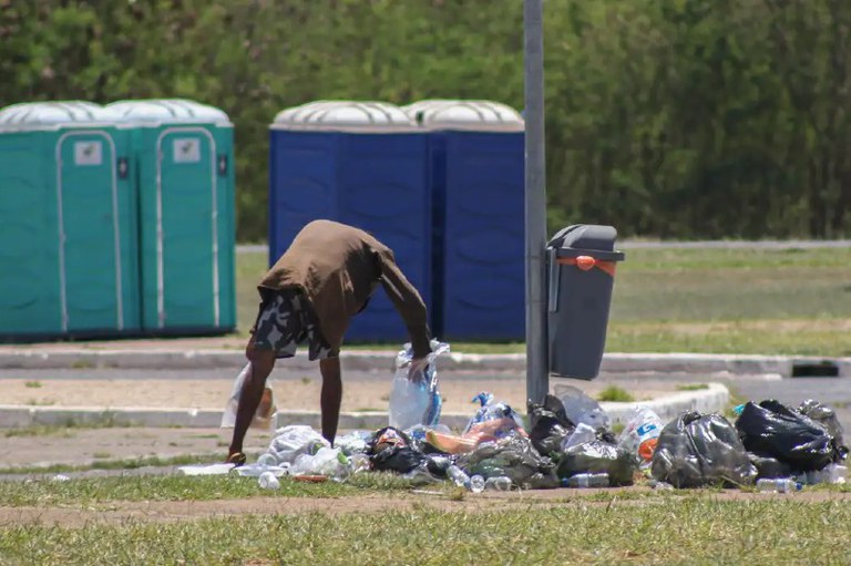 Foto mostra uma pessoa em situação de rua mexendo no lixo. Ela está de costas