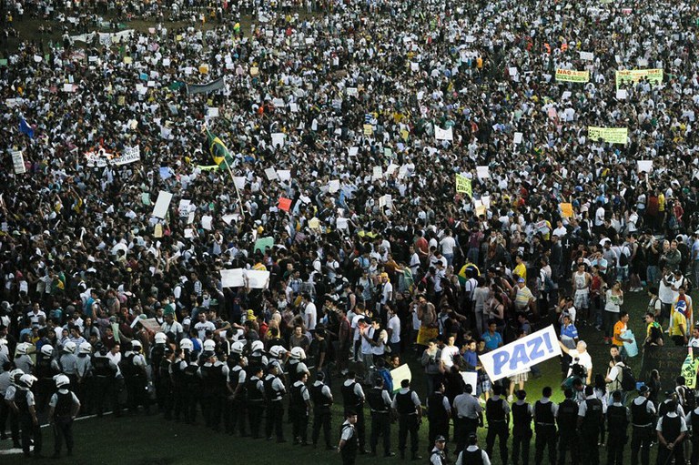 Foto mostra uma grande multidão protestando. Há policiais fazendo a segurança