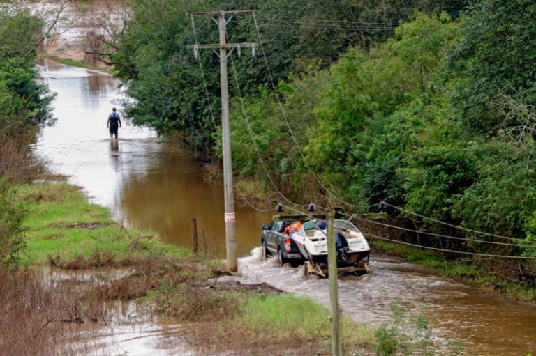 Foto de uma rua alagada com um carro passando pela água e árvores ao redor