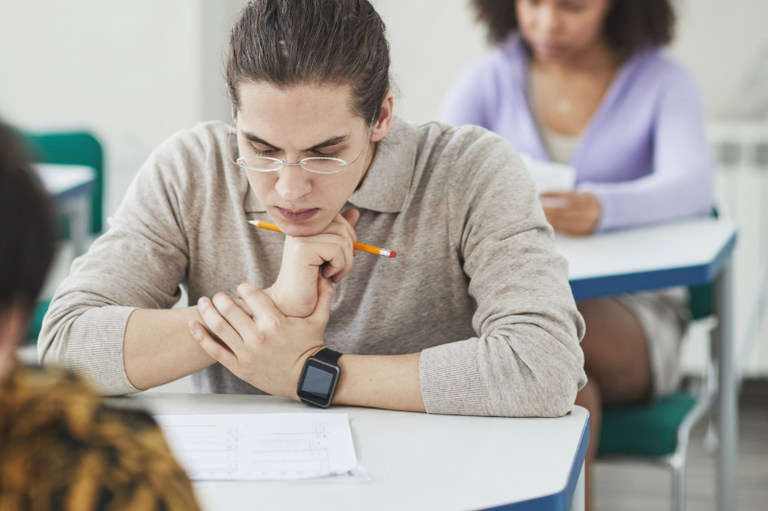 Um homem está sentado, olhando para um caderno de provas e pensativo. Atrás dele vemos uma mulher realizando a mesma prova. Ambos dentro de uma sala de aula.
