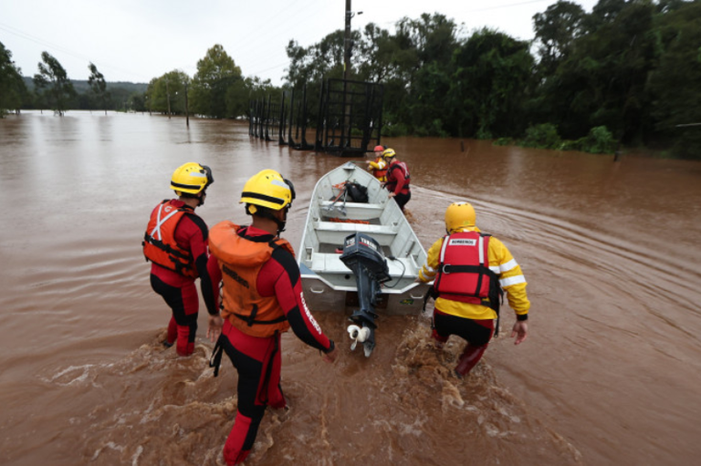 Fotografia de uma operação de resgate durante uma enchente urbana. Um bote inflável laranja, tripulado por três bombeiros com capacetes e coletes, navega por uma rua completamente submersa. No bote, estão vários civis e um cachorro resgatados. Ao fundo, casas e sobrados estão com a água na altura do primeiro andar, e a vegetação emerge parcialmente da inundação sob um céu nublado.
