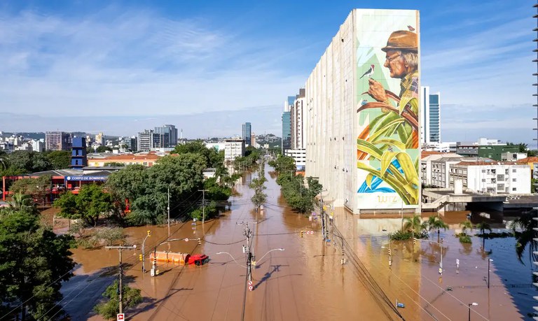 Fotografia tirada à distância mostra o centro de Porto Alegre alagado pelas enchentes de 2024. As ruas estão cobertas por água cor de lama, que atinge até o meio dos postes de iluminação. É possível ver também a boleia vermelha de um caminhão parcialmente submerso.