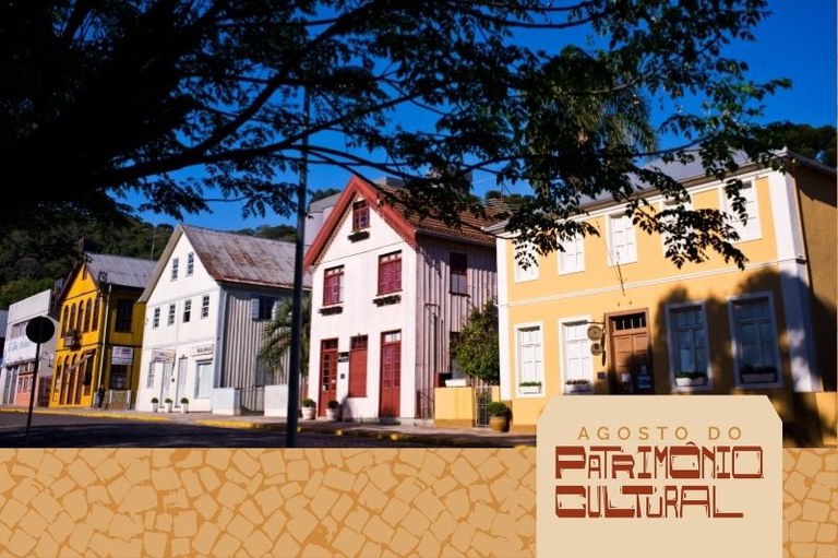 Fileira de casarões históricos e coloridos em uma rua, sob a sombra de árvores e céu azul. Na parte inferior, destaca-se um banner com a inscrição "Agosto do Patrimônio Cultural".