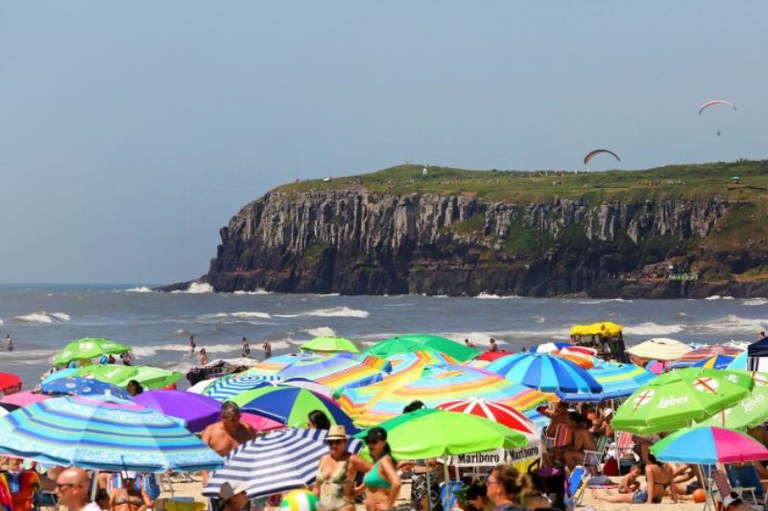 Dia de intensa movimentação de banhistas, em um dia ensolarado de verão, em uma praia do litoral norte gaúcho, com vários guarda-sóis, o mar e uma barreira de rochas ao fundo.