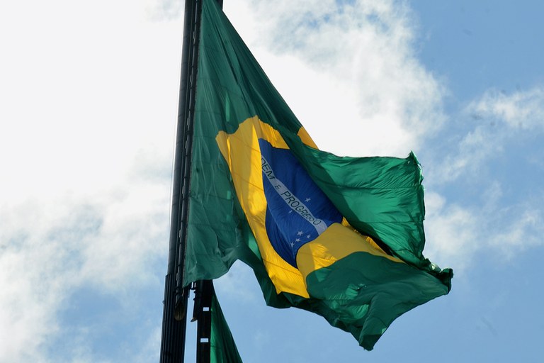 Bandeira brasileira tremulando com o céu azul ao fundo durante cerimônia da troca da bandeira em Brasília
