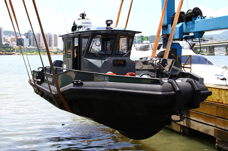 A embarcação adquirida pela Polícia Militar Ambiental, içada por quatro cordas de cada lado, flutua sobre a água de um cais. Ela é decorada com uma estampa de camuflagem e, no centro dela, há uma cabine envidraçada.