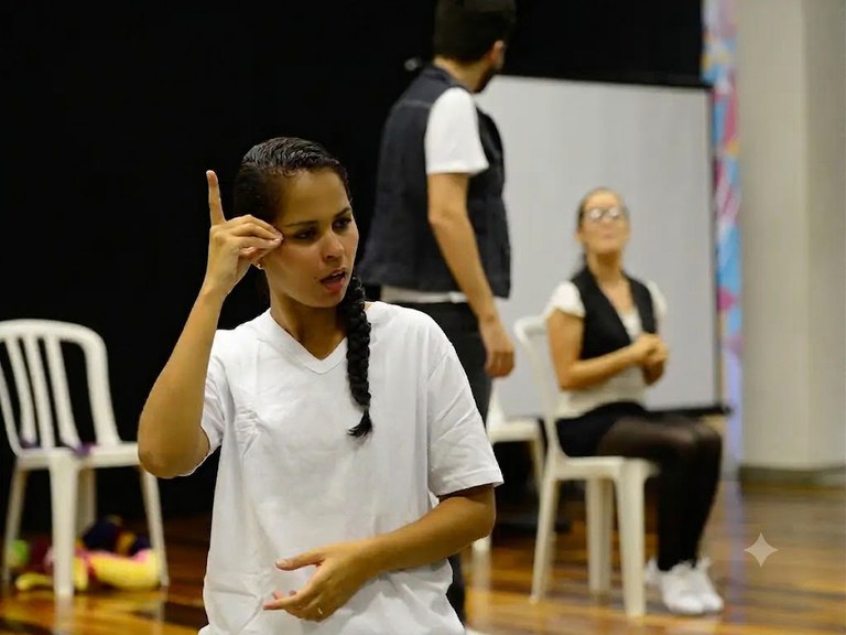 Uma mulher em primeiro plano faz um sinal em Libras vestindo uma camiseta branca e lisa. Ao fundo, um homem de pé e outra mulher sentada em uma cadeira observam a cena.
