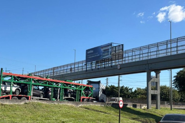 Foto da passarela na BR101 com um painel publicitário e um caminhão cegonheiro passando por baixo
