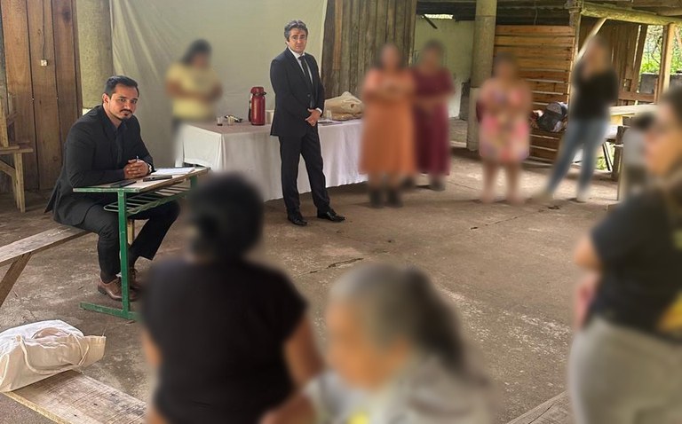 Foto de uma reunião em um espaço semiaberto e rústico (possivelmente uma aldeia ou galpão), com um grupo de mulheres indígenas e três homens, dois deles vestidos formalmente de terno. As mulheres estão em pé ou sentadas em bancos de madeira. A cena é focada em uma interação e diálogo entre os grupos.