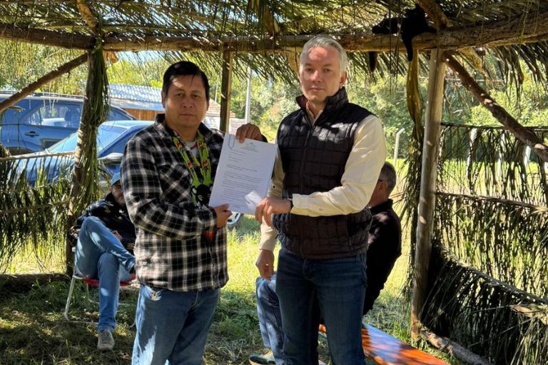 Dois homens em pé seguram juntos um documento em papel sob uma estrutura rústica com cobertura de palha. O homem à esquerda veste camisa xadrez e um colar indígena tradicional, e o homem à direita usa um colete escuro sobre camisa clara e calça jeans. Ao fundo, em ambiente externo, há vegetação, carros estacionados e outras pessoas sentadas.