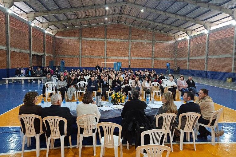 Foto mostra os participantes da mesa principal da audiência pública, vistos de costas. À frente deles, o público da audiência sentado em cadeiras plásticas. Ao lado e acima, veem-se as paredes e o teto do ginásio onde foi realizada a audiência.