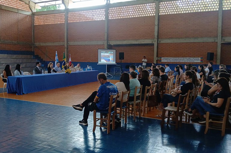 Foto mostra audiência pública ocorrendo com a população disposta em cadeiras escutando os participantes da mesa