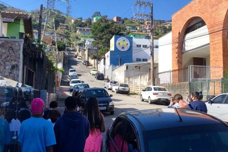 foto mostra uma rua do Morro do Mocotó em ladeira com carros estacionados ao longo do meio fio dos dois lados da rua, construções em cada lado da rua e pessoas andando