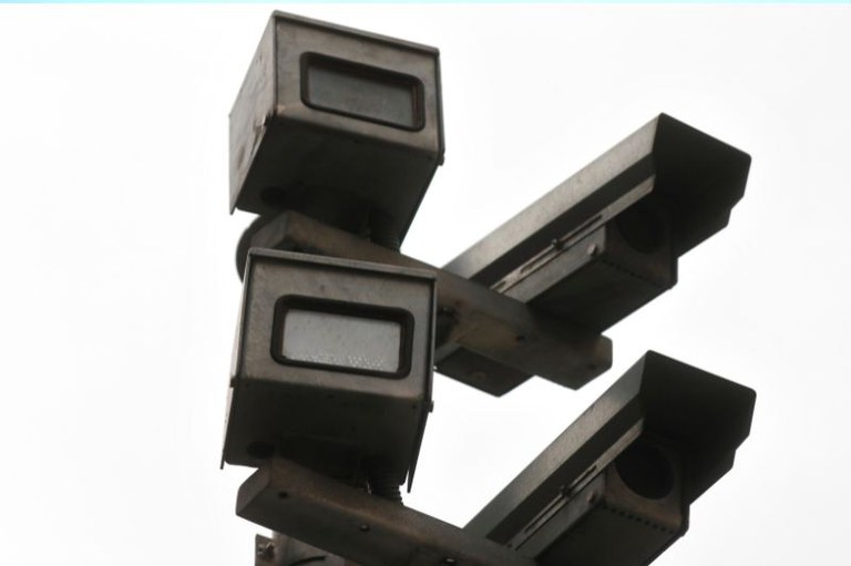 Foto mostra um conjunto de radares num post, contra um fundo claro.