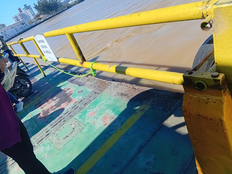 Fotografia tirada no interior de um ferry boat mostra as barras amarelas de proteção para evitar que pessoas caiam no mar. Há um grande vão entre as barras. Ao fundo, o mar e a cidade.