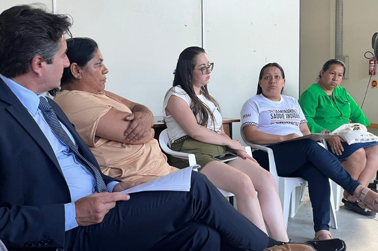 Foto mostra quatro mulheres e um homem de paletó sentados em cadeiras de plástico em ambiente interno