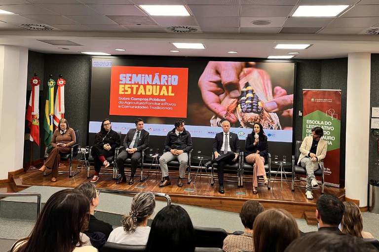 Foto no auditório do evento mostra sete pessoas entre homens e mulheres usando roupas formais sentadas em cadeiras de frente para o público que acompanha o seminário. ao fundo, um telão com o nome do evento Seminário Estadual - sobre compras públicas da agricultura familiar e povos e comunidades tradicionais