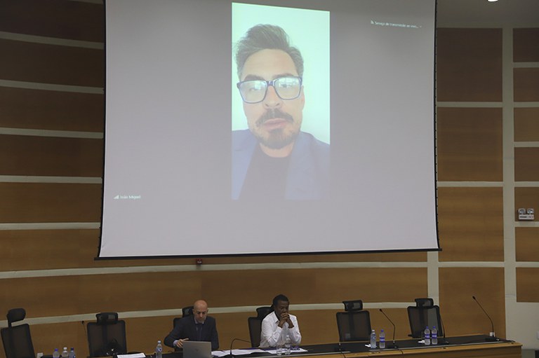Duas pessoas sentadas à mesa dos palestrantes de um auditório. Acima delas, em um telão, a imagem de uma terceira pessoa.