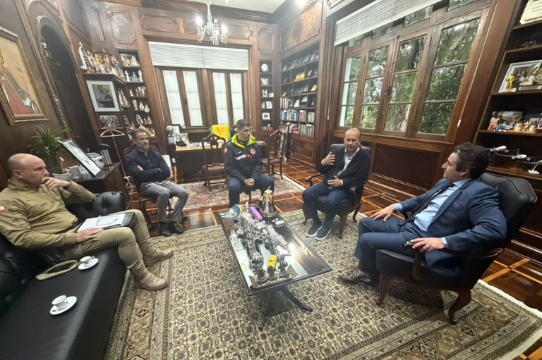 Cinco homens, incluindo autoridades civis e militares fardados, reunidos em um escritório clássico com revestimento de madeira, estantes de livros e tapete ornamentado.