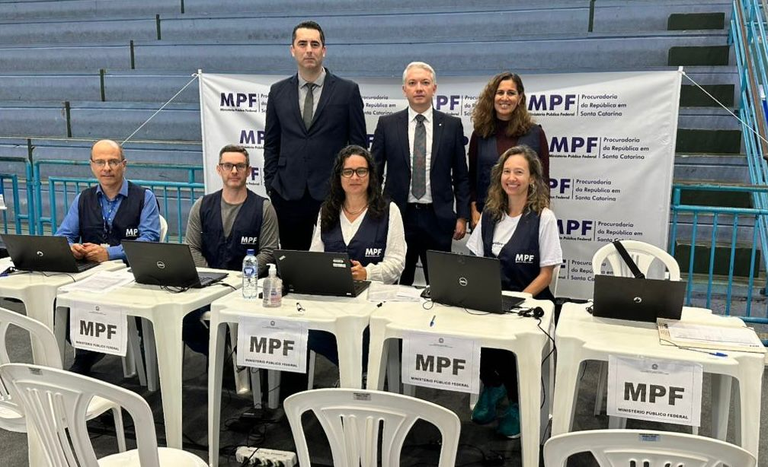 Dois homens vestidos de terno e uma mulher vestida de preto em pé, dois homens e duas mulheres vestindo coletes do MPF e sentados em uma mesa com laptops em cima, vestindo coletes do MPF. Ao fundo um banner com várias logos do Ministério Público Federal.