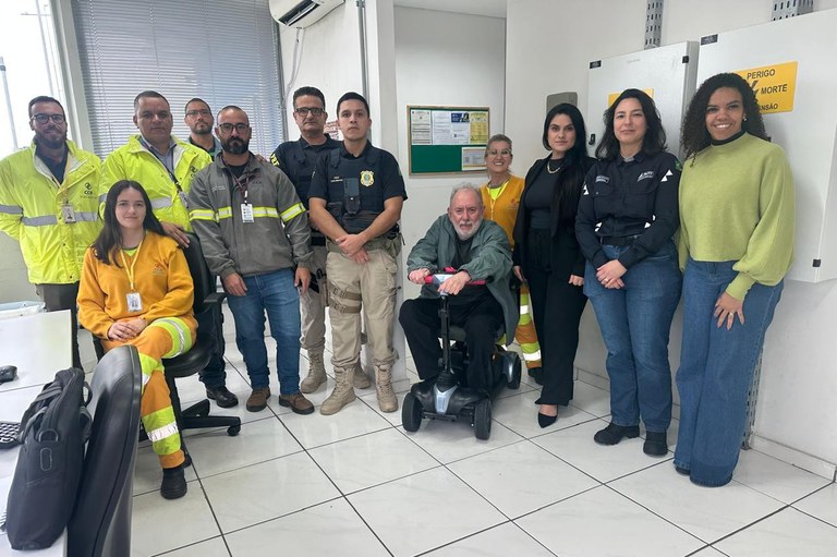 Foto de comunicação do MPF sobre a fiscalização de um posto de pesagem na BR-101 em Araranguá, SC, que mostra um grupo diverso de aproximadamente doze pessoas reunidas em um ambiente interno, que se assemelha a um escritório ou sala de controle. O grupo é composto por homens e mulheres, com alguns vestindo coletes de segurança nas cores amarelo neon e cinza (provavelmente representando a concessionária da rodovia, como a CCR, e/ou equipes técnicas), um homem ao centro em uniforme da Polícia Rodoviária Federal (PRF), um homem mais velho sentado em uma cadeira de rodas motorizada, e algumas mulheres em trajes sociais ou casuais, à direita. A diversidade de vestimentas sugere a participação de diferentes órgãos e entidades na ação de fiscalização, como o Ministério Público Federal (MPF), agentes de segurança e possivelmente a equipe da concessionária da rodovia.