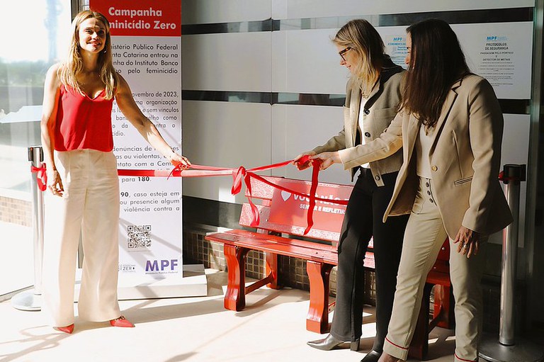A procuradora Rafaella Alberici, à esquerda, e as procuradoras Patrícia Muxfeldt e Jerusa Viecili, à direita, seguram a fita inaugural do banco vermelho, que está atrás delas. À esquerda, um totem exibe informações sobre a campanha Feminicídio Zero.