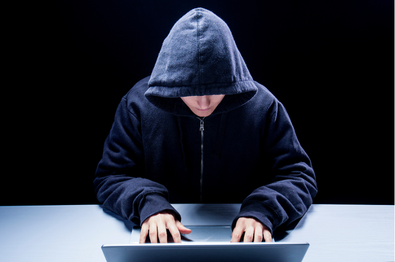 Foto mostra homem com a cabeça abaixada vestido com moletom azul escuro, com capuz, sentado à mesa com as mãos sobre teclado de notebook.