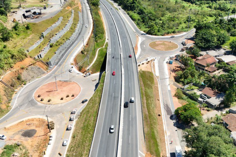 Vista aérea de um trecho duplicado de uma rodovia asfaltada com veículos em movimento, integrada a duas rotatórias laterais e cercada por áreas verdes e residências.