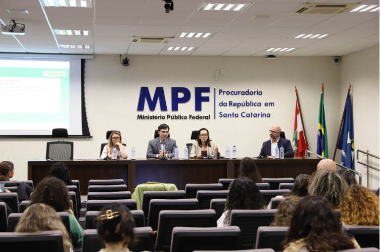 Ao fundo de um auditório, quatro pessoas sentadas à mesa de abertura do evento falam para uma plateia em primeiro plano, vista de costas. Na parede ao fundo, a logomarca do MPF em Santa Catarina. À direita, as bandeiras do estado de Santa Catarina, do Brasil e do Ministério Público.