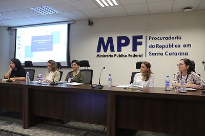 Na mesa dos palestrantes, cinco mulheres sentadas lado a lado. Ao fundo, uma tela em que se projeta uma imagem e a logomarca do MPF.