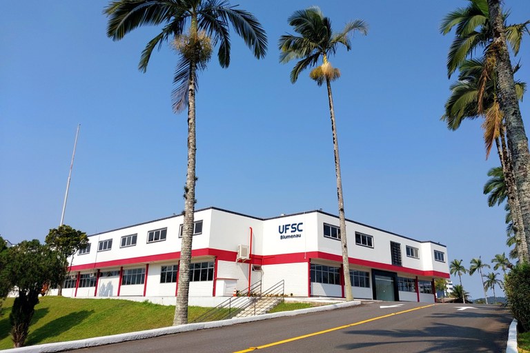 A imagem apresenta uma vista externa do campus da UFSC (Universidade Federal de Santa Catarina) em Blumenau, sob um céu azul limpo e ensolarado. O edifício principal é uma construção moderna de dois pavimentos com fachada branca, destacada por uma faixa horizontal vermelha vibrante que percorre toda a extensão da estrutura e janelas retangulares organizadas de forma simétrica. Em primeiro plano, uma estrada asfaltada com sinalização viária sobe suavemente em direção à entrada, ladeada por um gramado verde bem cuidado e três palmeiras imperiais altas e esguias que emolduram a cena. À esquerda, o logotipo "UFSC Blumenau" está estampado na parede branca, identificando a instituição em um ambiente que transmite uma sensação de organização e tranquilidade acadêmica.