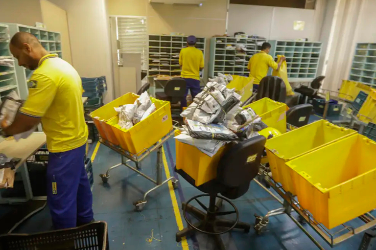 Foto mostra carteiros trabalhando em uma unidade de distribuição de cartas e encomendas