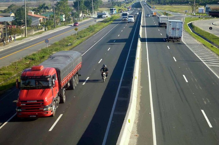 Vista superior de uma rodovia duplicada com mureta central, destacando um caminhão vermelho e uma motocicleta em primeiro plano.