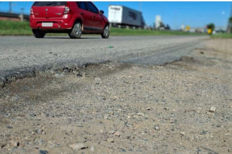 Fotografia em close do asfalto desgastado e com buracos em uma via marginal. Ao fundo, em plano desfocado, um carro vermelho e um caminhão trafegam sob céu azul.