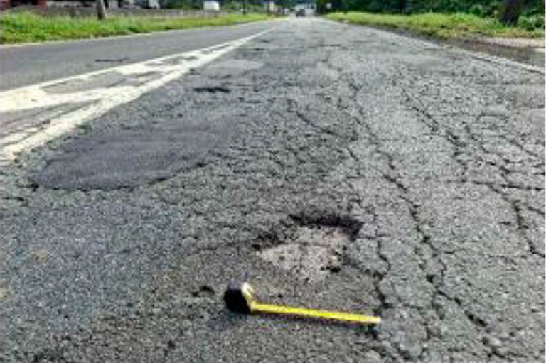 A imagem mostra trecho da BR-101 com asfalto cedendo, buracos e falta de manutenção básica. Perícia técnica do MPF utilizou instrumentos de medição para documentar a situação da rodovia