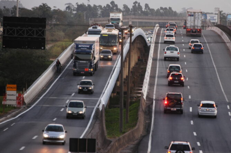 Foto mostra tráfego intenso de caminhões e carros em uma rodovia de pista dupla em uma ponte ou viaduto, ao entardecer.