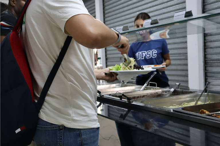 Foto mostra estudantes se servindo no RU do Campus. Uma estudante veste uma blusa azul onde está escrito UFSC