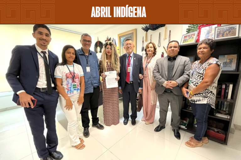 A imagem mostra um grupo de oito pessoas posando juntas em um ambiente interno bem iluminado. No centro, uma mulher veste uma roupa estampada, segurando um papel. Ao redor dela estão homens e mulheres, alguns de terno e outros com roupas casuais. Atrás, há uma parede decorada com artesanato indígena, incluindo cocares e objetos culturais, além de prateleiras com quadros e livros.