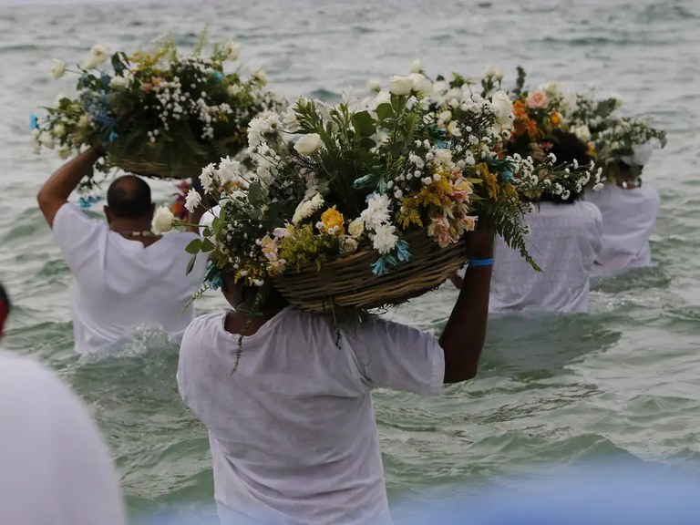 Pessoas vestidas com roupas brancas caminham dentro do mar carregando cestos ornamentados com flores coloridas sobre a cabeça. A cena retrata uma oferenda tradicional em celebração de tradição afro-brasileira (como a homenagem a Iemanjá).