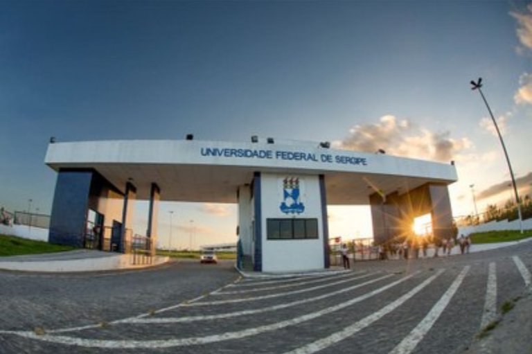 Foto da entrada do campus da Universidade Federal de Sergipe em Lagarto em tarde de céu azul, com o sol a se pôr