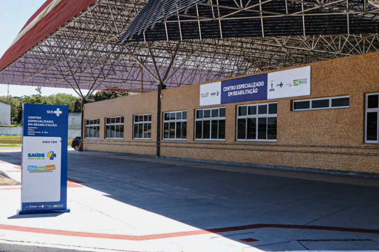 A imagem mostra a fachada de um prédio térreo, de tijolos aparentes, com grandes janelas retangulares e um amplo toldo metálico cobrindo a entrada. Em destaque, há uma placa azul e branca na calçada, indicando “Centro Especializado em Reabilitação” e o logotipo do SUS. Acima da porta principal, outra placa exibe o mesmo nome da unidade, além de logotipos de órgãos públicos. O espaço externo é amplo e acessível, com piso liso e sinalização no chão.
