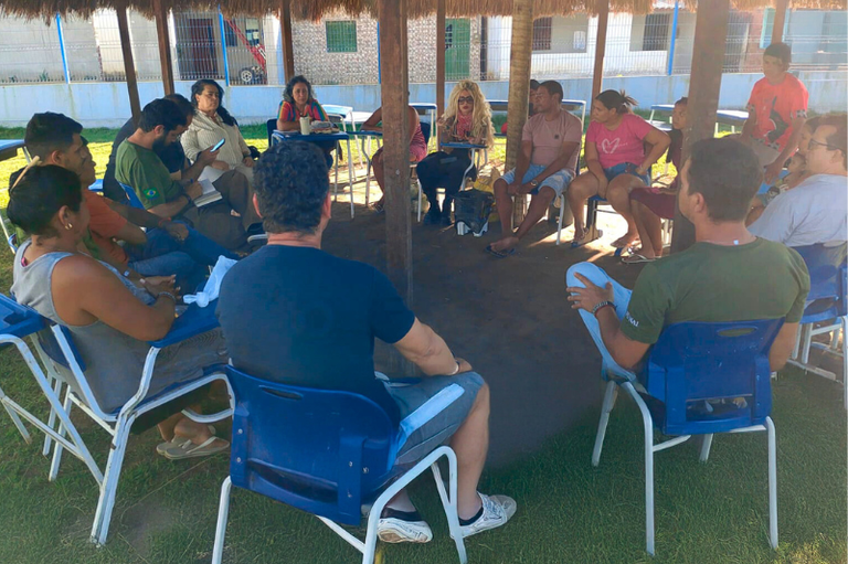 Um grupo de pessoas sentadas em círculo, conversando durante uma reunião. Eles estão sob uma estrutura que parece um quiosque, com telhado de palha.