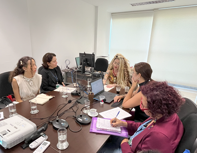 A imagem mostra uma reunião com seis participantes sentados em uma mesa de trabalho. No centro, há um laptop aberto e acessórios de apresentação como projetores. As pessoas estão atentas, algumas tomando notas. À esquerda, uma mulher de cabelo encaracolado e vestido claro observa. Ao centro, uma mulher com cabelos longos, vestindo uma blusa colorida, fala enquanto é ouvida atentamente. À direita, duas mulheres conversam: uma com cabelo cacheado e blusa roxa, e outra com cabelo curto e colorido, usando um blazer. A atmosfera parece ser de colaboração e discussão, em um ambiente bem iluminado.