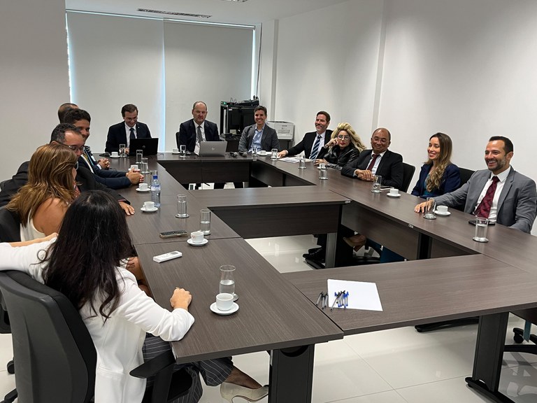 Fotografia em ambiente interno e bem iluminado mostra doze pessoas, vestidas formalmente em trajes profissionais, sentadas ao redor de uma grande mesa de reunião marrom-escura em formato de U. O grupo, composto por homens e mulheres dispostos ao longo dos lados da mesa, parece engajado em uma reunião de trabalho, com copos, xícaras de café, papéis e notebooks dispostos à sua frente sobre o piso de porcelanato claro.