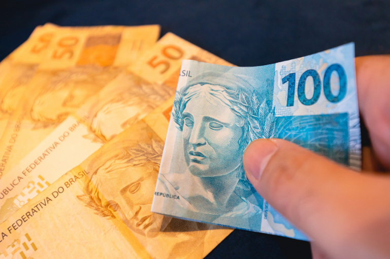 Um close-up de uma mão segurando uma nota de R$ 100 reais (azul) dobrada. No fundo, há um punhado de notas de R$ 50 reais (amarelas) espalhadas.