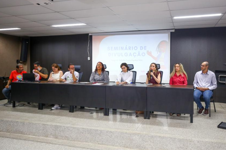 A imagem mostra uma mesa de conferência com nove pessoas sentadas em um auditório. Ao fundo, há um telão projetando um slide com o título ¿SEMINÁRIO DE DIVULGAÇÃO¿. A sala tem paredes escuras e iluminação branca no teto. As pessoas na mesa vestem roupas formais e casuais. Uma mulher ao centro fala ao microfone, enquanto os demais aparentam ouvir ou tomar notas. Alguns usam camisetas brancas com logos, outros roupas sociais. A mesa preta é longa e tem cadeiras pretas. O ambiente sugere um evento acadêmico ou institucional.