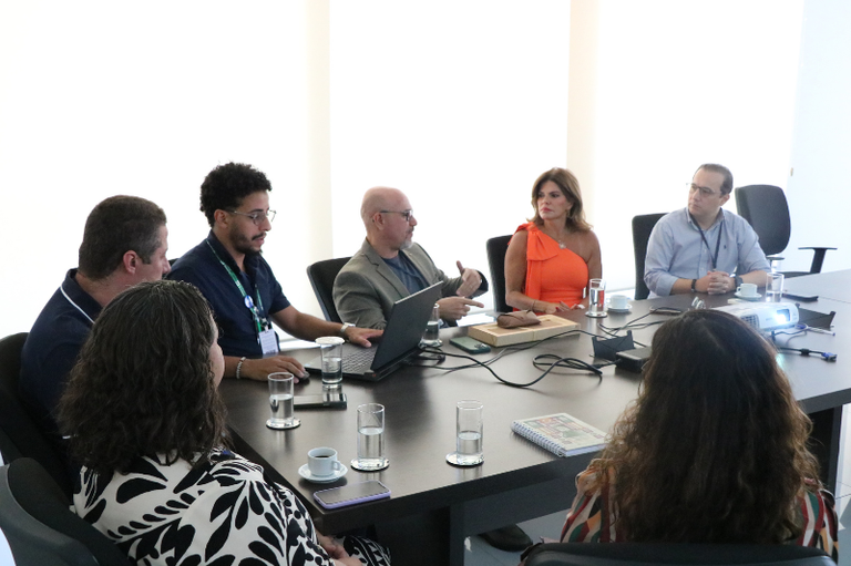 Sete pessoas estão reunidas em torno de uma mesa de conferência escura em uma sala bem iluminada. No centro, um homem de paletó cinza gesticula enquanto fala, posicionado em frente a um laptop aberto. Ao seu lado, uma mulher veste um vestido laranja vibrante de ombro único. Sobre a mesa, há copos de água, xícaras de café, um projetor e cadernos. Duas mulheres são vistas de costas em primeiro plano, completando o círculo da reunião.