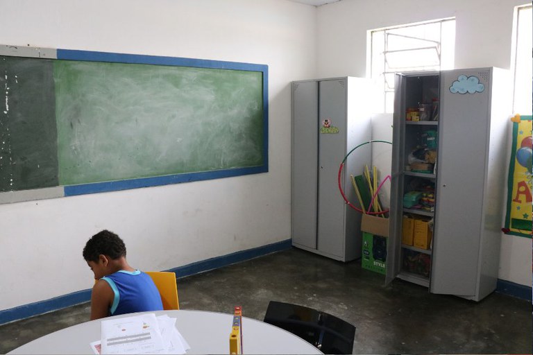 Foto mostra uma sala de aula, com um quadro de giz verde, cadeiras e uma criança sentada de cabeça baixa virada para o quadro