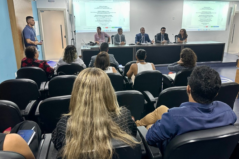 A imagem mostra uma sala de conferências onde um grupo de pessoas participa de uma reunião. No centro, há uma mesa com cinco pessoas sentadas, vestidas formalmente, com microfones e garrafas d'água. Atrás delas, duas telas projetam um documento. No público, várias pessoas estão sentadas em cadeiras pretas, algumas tomando notas. À esquerda, um homem de camisa azul está de pé, segurando um celular. O ambiente tem paredes azuis e brancas, com iluminação clara e moderna.