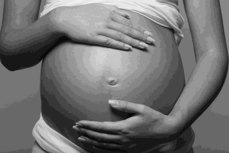Imagem em preto e branco de uma barriga de mulher grávida, vista frontal. A mulher está com as duas mãos posicionadas sobre a barriga.